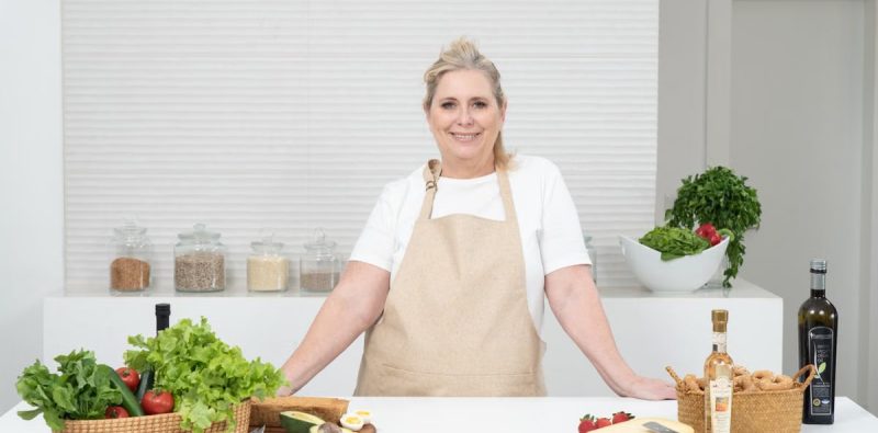A mature woman in an apron stands at a kitchen counter with fresh produce and cheese, ready for cooking.